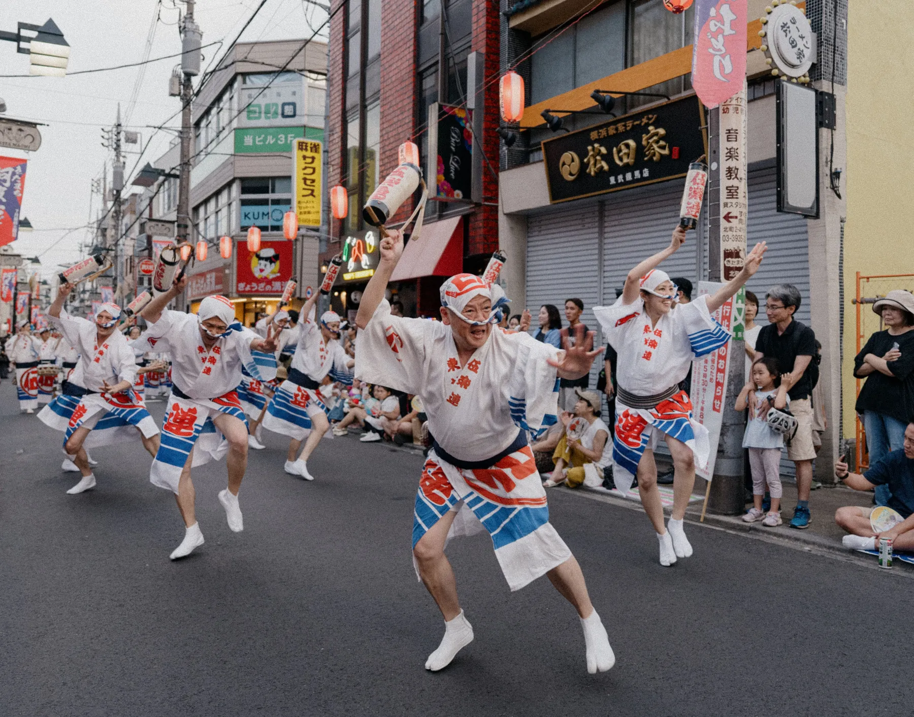Dancers in white and blue happi coats move down a closed street at a Japanese festival while onlookers watch from the kerb